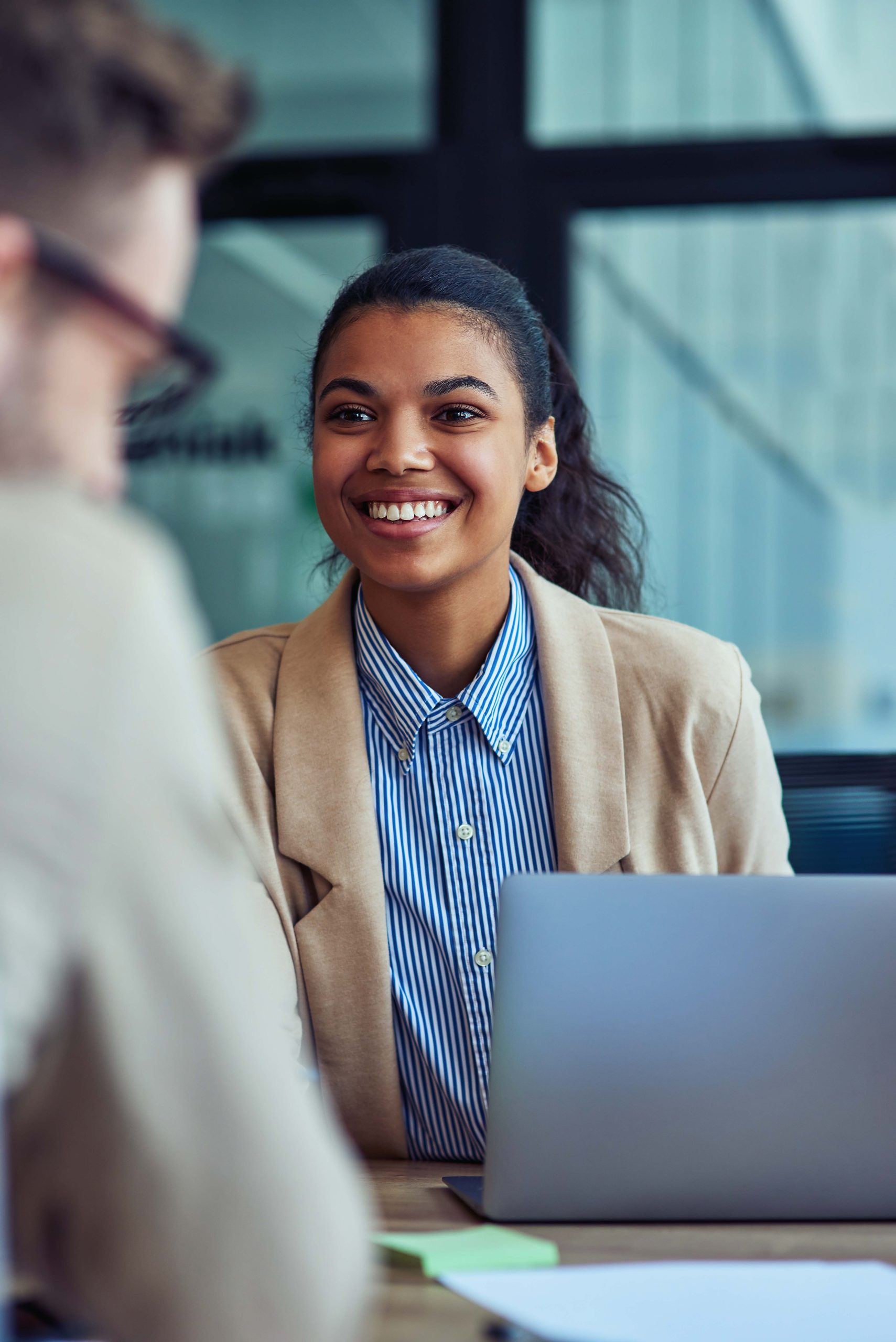 Vertical shot of a young cheerful mixed race female office worker working on laptop in the modern office, talking with colleague and smiling